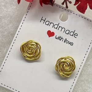 May include: A pair of gold-colored wire earrings shaped like roses. The earrings are on a white card with the text "Handmade with love".