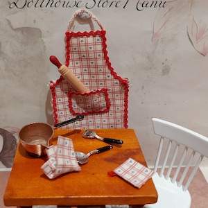 Op de afbeelding: Een miniatuur keukenset voor poppenhuizen met een rood-wit geblokte schort, een houten deegroller, een koperen pan en een witte stoel. De set staat op een houten tafel met een geblokt tafelkleed.