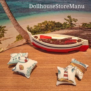 Può includere: Quattro cuscini in miniatura con tema nautico, con fari, conchiglie e salvagente. I cuscini sono disposti su una superficie di legno accanto a una barca in miniatura rossa e bianca con una rete da pesca marrone.