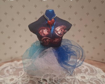Puede incluir: Una forma de vestido en miniatura para casa de muñecas con una falda de tul azul y blanco, un corpiño de cinta azul y roja y un collar de corazón rojo. La forma de vestido está sobre un encaje de encaje blanco.