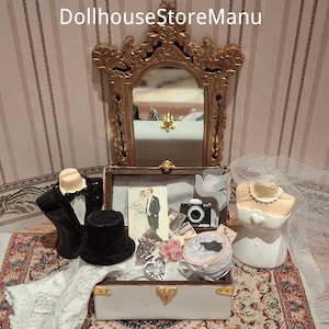 May include: A white wooden chest with a gold latch and hinges. The chest is open and contains a variety of miniature wedding-themed items, including a black tuxedo, a top hat, a white dress, a veil, a camera, and a pair of doves. The chest is sitting on a patterned rug.