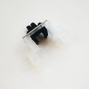 May include: A small, plastic component with a black central piece and translucent white extensions. A thin, silver-colored metal strip is attached across the top. The object appears to be a part of a larger mechanism or device.