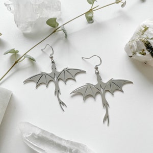 May include: A pair of silver dragon earrings with delicate wings and a detailed tail. The earrings are hanging from silver hooks.