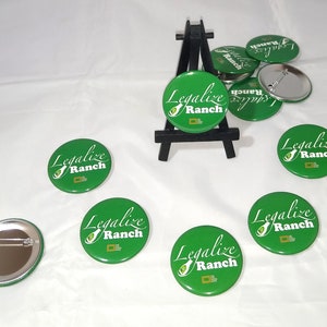 Puede incluir: Ocho botones redondos verdes con el texto "Legalize Ranch" en letras blancas. Los botones tienen una imagen blanca de un aguacate con una botella de aderezo ranch dentro. Los botones están sobre una superficie blanca con un pequeño caballete de madera.