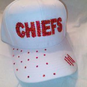 May include: White baseball cap with the word "CHIEFS" in red rhinestones across the front. The cap has additional red rhinestones scattered on the brim and a small cluster of red rhinestones on the side.