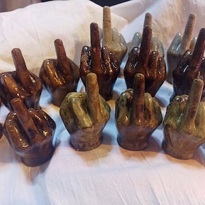 May include: A collection of ceramic hand figurines, each with a middle finger raised in a gesture of defiance. The figurines are painted in various shades of brown, green, and gray, with some featuring a textured finish.