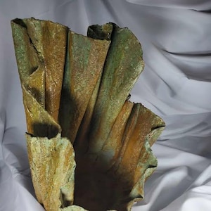 May include: A textured, abstract sculpture with a folded, organic form. The piece features a blend of earthy tones, including shades of brown, green, and gold, creating a weathered appearance. The open interior suggests a vessel or decorative object.