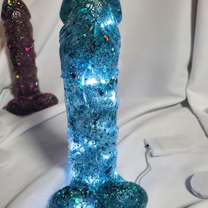 May include: A blue and teal glitter-covered sculpture with internal lights. The sculpture is shaped like a phallus. A second, smaller purple sculpture is visible in the background. The sculpture is on a white surface.