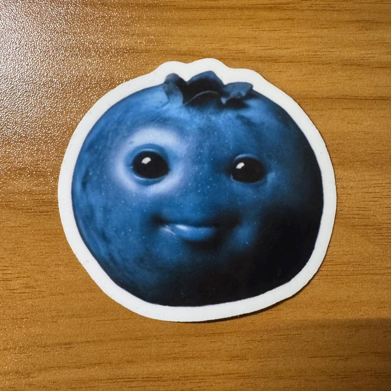 Puede incluir: Una pegatina con una caricatura de ar&aacute;ndano con una cara sonriente. El ar&aacute;ndano es de color azul oscuro con un borde blanco. El ar&aacute;ndano tiene ojos grandes y negros y una peque&ntilde;a lengua que sobresale.