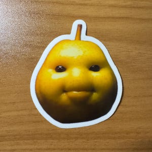 Puede incluir: Una pegatina de pera amarilla con un dise&ntilde;o de cara sonriente. La pera tiene ojos oscuros y una boca peque&ntilde;a y sonriente. La pegatina est&aacute; delineada en blanco y est&aacute; sobre una superficie de madera.