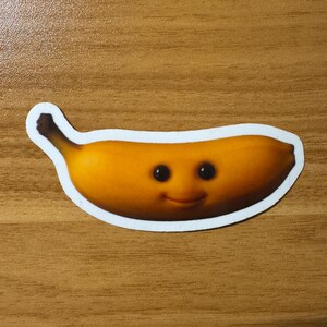 Puede incluir: Una pegatina con un pl&aacute;tano de dibujos animados sonriente con ojos negros y una expresi&oacute;n feliz. El pl&aacute;tano es amarillo con sombras marrones y un borde blanco. La pegatina est&aacute; sobre una superficie de madera.