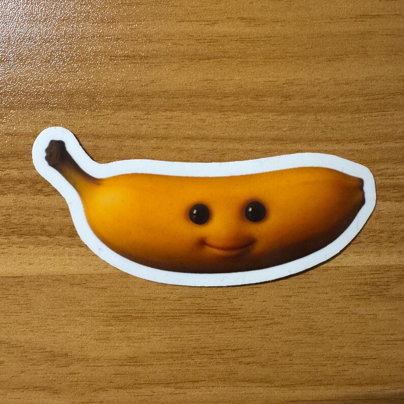 Puede incluir: Una pegatina con un pl&aacute;tano de dibujos animados sonriente con ojos negros y una expresi&oacute;n feliz. El pl&aacute;tano es amarillo con sombras marrones y un borde blanco. La pegatina est&aacute; sobre una superficie de madera.