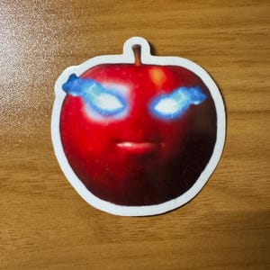 Puede incluir: Una pegatina con una manzana roja con una cara. La manzana tiene ojos azules brillantes y una boca peque&ntilde;a. La pegatina est&aacute; sobre una superficie de madera.