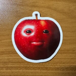 Puede incluir: Una pegatina de una manzana roja con una cara sonriente. La pegatina tiene un borde blanco y un peque&ntilde;o tallo en la parte superior. La cara de la manzana tiene dos ojos negros y una boca peque&ntilde;a y curvada.