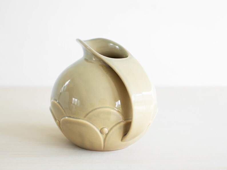 May include: A light green ceramic pitcher with a rounded body and a curved handle. The pitcher has a decorative design of raised circles and semi-circles.