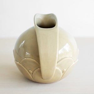 May include: A light green ceramic pitcher with a rounded body and a curved spout. The pitcher has a decorative design of overlapping circles.