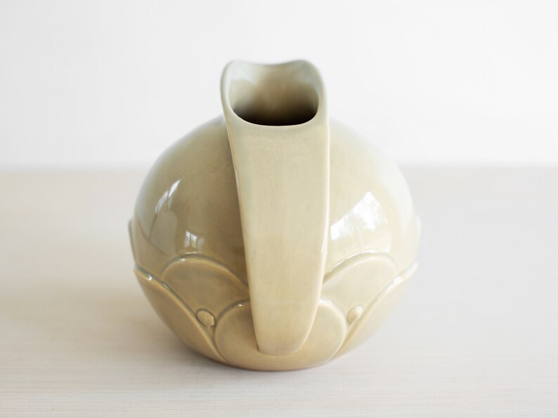 May include: A light green ceramic pitcher with a rounded body and a curved spout. The pitcher has a decorative design of overlapping circles.