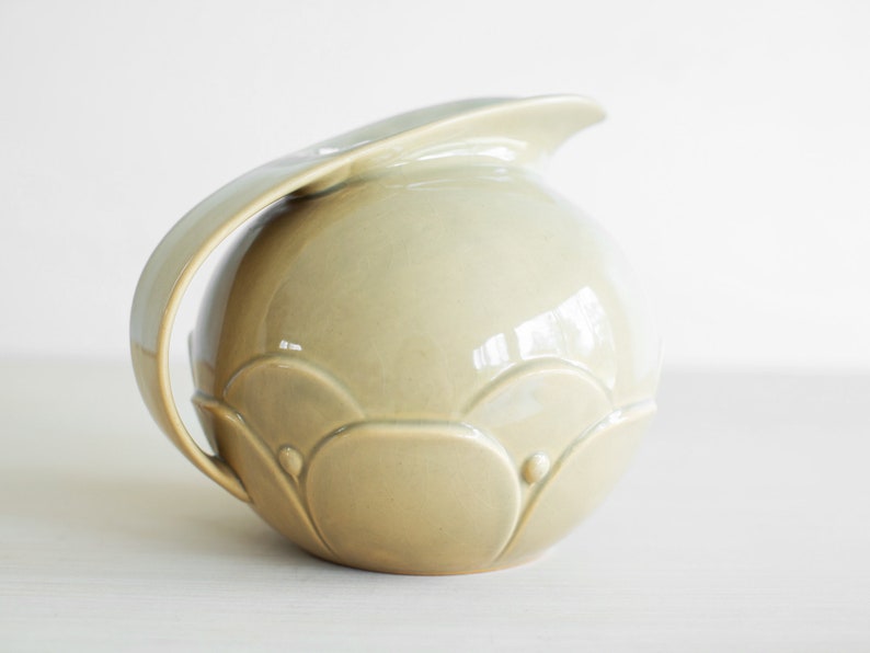 May include: A light green ceramic pitcher with a rounded body and a curved handle. The pitcher has a decorative design of raised circles and scalloped edges.