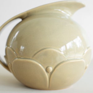 May include: A light beige ceramic pitcher with a rounded body and a curved handle. The pitcher has a decorative design of raised flower petals around the bottom.