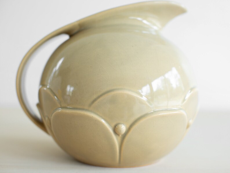 May include: A light beige ceramic pitcher with a rounded body and a curved handle. The pitcher has a decorative design of raised flower petals around the bottom.
