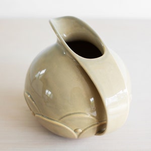 May include: A light green ceramic pitcher with a unique, curved design. The pitcher has a smooth, glossy finish and a wide opening.