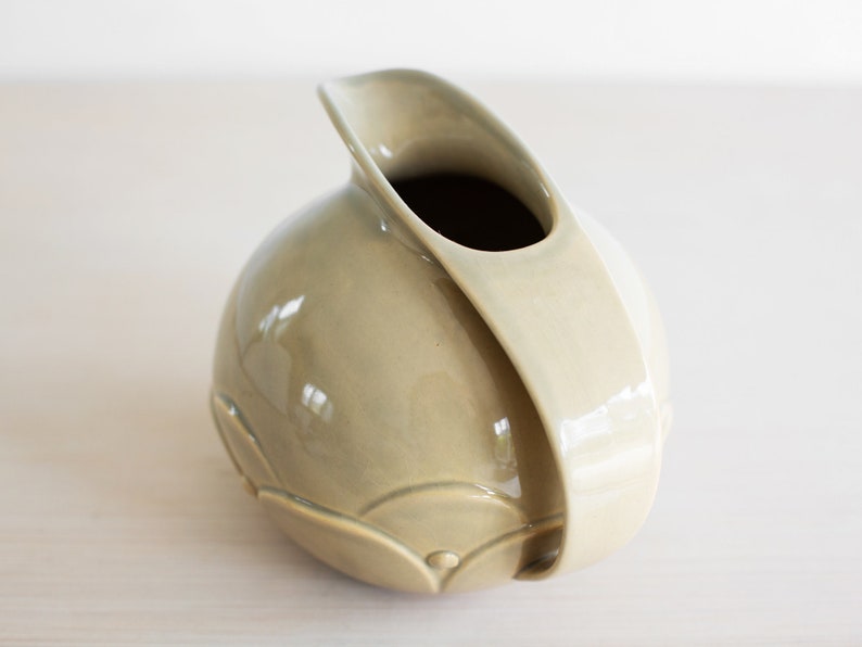 May include: A light green ceramic pitcher with a unique, curved design. The pitcher has a smooth, glossy finish and a wide opening.