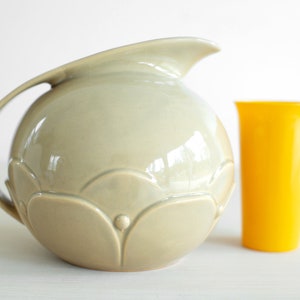May include: A light gray ceramic pitcher with a rounded body and a decorative floral design. The pitcher has a wide mouth and a handle. It is sitting on a white surface next to a yellow plastic cup.