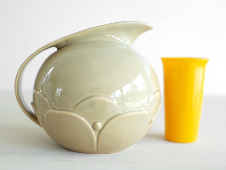May include: A light gray ceramic pitcher with a rounded body and a decorative floral design. The pitcher has a wide mouth and a handle. It is sitting on a white surface next to a yellow plastic cup.