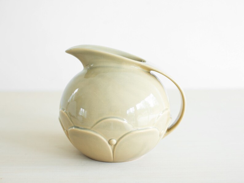 May include: A light green ceramic pitcher with a rounded body and a curved handle. The pitcher has a textured surface with a floral design.