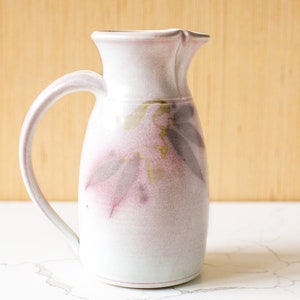 May include: A white ceramic pitcher with a floral design in shades of pink and green. The pitcher has a handle and a spout.