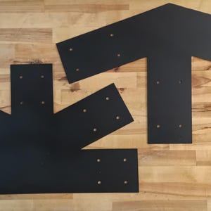 May include: Two matte black metal structural plates for timber framing. One plate is T-shaped with a pointed top, and the other is a multi-angled connector plate with several pre-drilled circular holes for mounting hardware on both pieces.