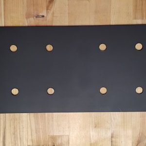 May include: A black metal plate with six evenly spaced holes. The plate has rounded edges and a slightly textured surface.
