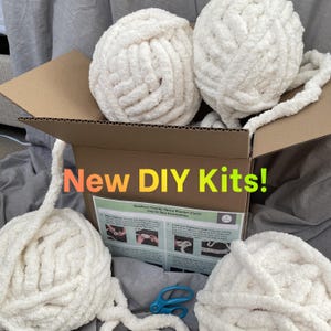 Pode incluir: Uma caixa de cartão cheia de bolas de fio branco grandes para um kit de manta grossa DIY. A caixa também contém instruções e uma tesoura azul. O texto na caixa diz "New DIY Kits!"