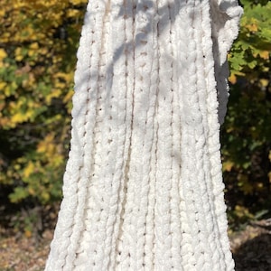 May include: A white chunky knit throw blanket with a soft, fuzzy texture. The blanket is draped over a wooden stick and is hanging in front of a blurred background of fall foliage.