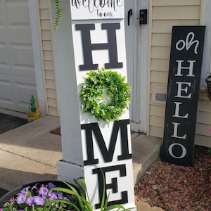 May include: A white wooden sign with black letters spelling "HOME" with a green wreath between the "H" and "M". The sign is leaning against a white post. A black sign with white letters spelling "OH HELLO" is leaning against a yellow wall.