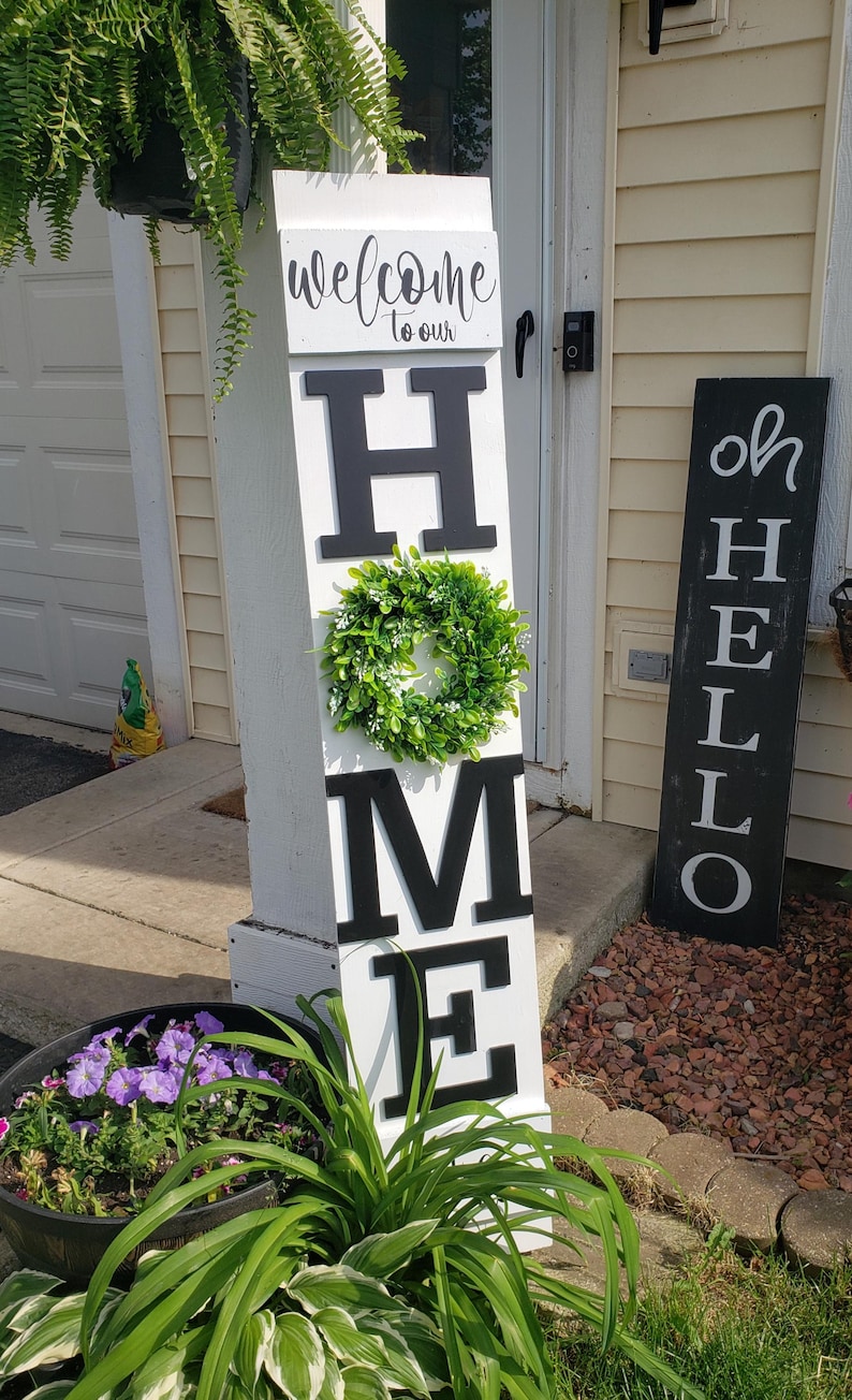 May include: A white wooden sign with black letters spelling "HOME" with a green wreath between the "H" and "M". The sign is leaning against a white post. A black sign with white letters spelling "OH HELLO" is leaning against a yellow wall.
