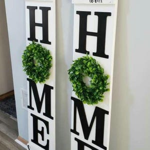 May include: Two white wooden signs with the words "welcome to our" and "Sweet home" in script. Large black letters spell "HOME" with a green wreath on each sign. Home decor.