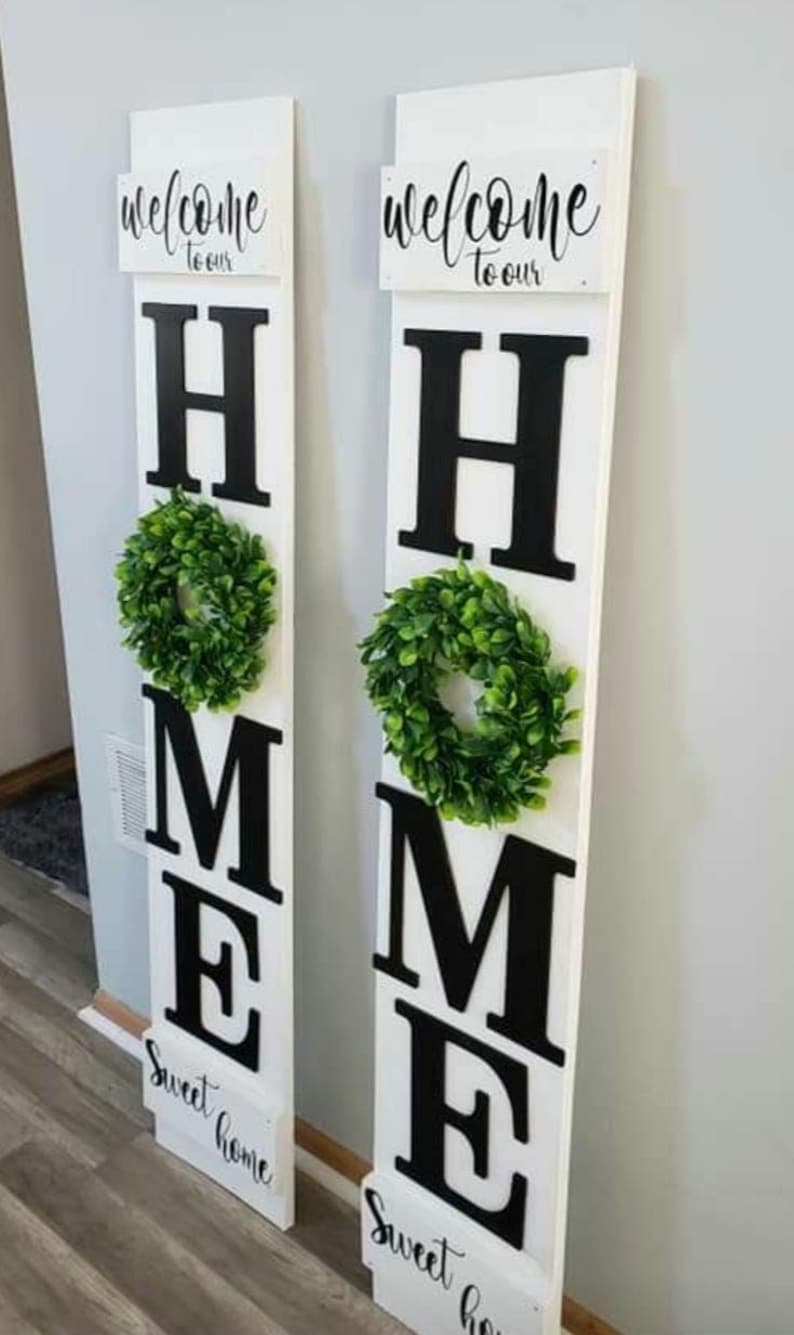 May include: Two white wooden signs with the words "welcome to our" and "Sweet home" in script. Large black letters spell "HOME" with a green wreath on each sign. Home decor.