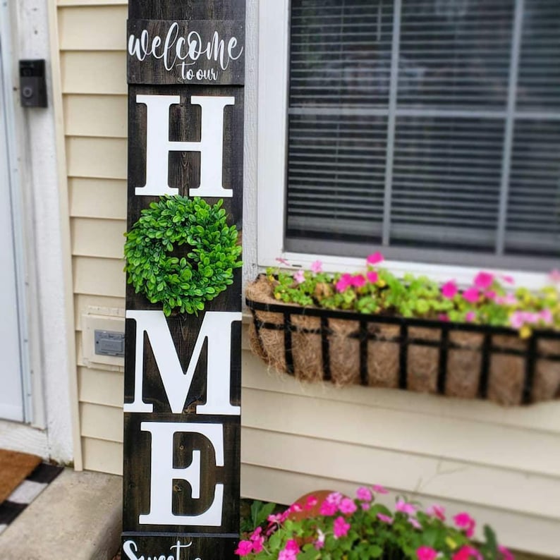 May include: A wooden welcome sign with the word "HOME" in large white letters. A green wreath is centered between the "H" and "M". The top of the sign reads "welcome to our" in cursive. The sign is placed outside a house.