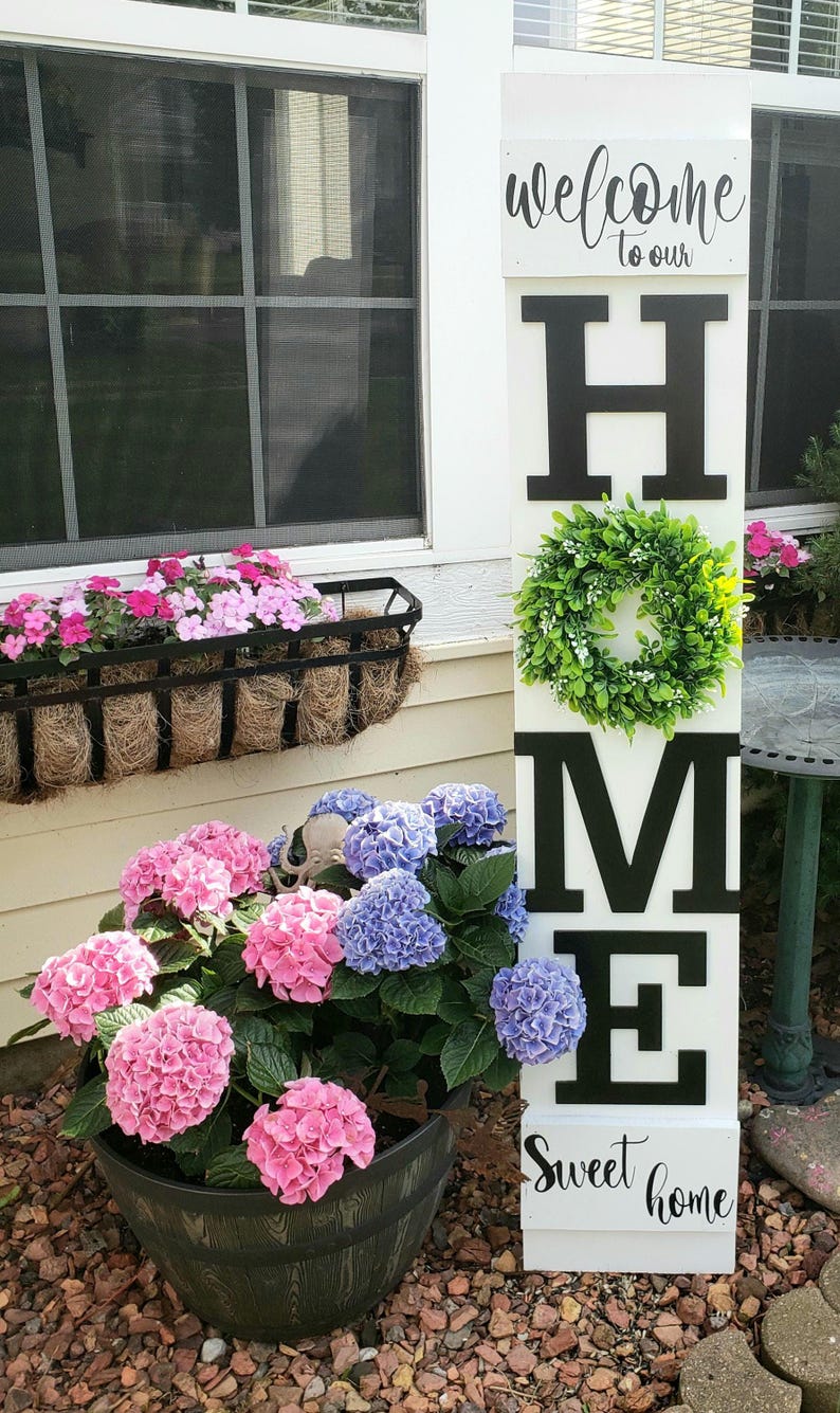 May include: A white wooden sign with the word "WELCOME" at the top and "HOME" in large black letters. A green wreath is centered on the "M". The sign also says "Sweet home". Pink and blue hydrangeas are in a dark pot.