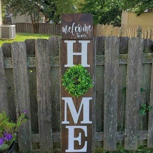 May include: A wooden sign with the word "HOME" in large white letters. The letter "O" is replaced with a green wreath. The sign also includes the phrases "welcome to our" and "Sweet home" in cursive script. The sign is placed in a grassy yard.