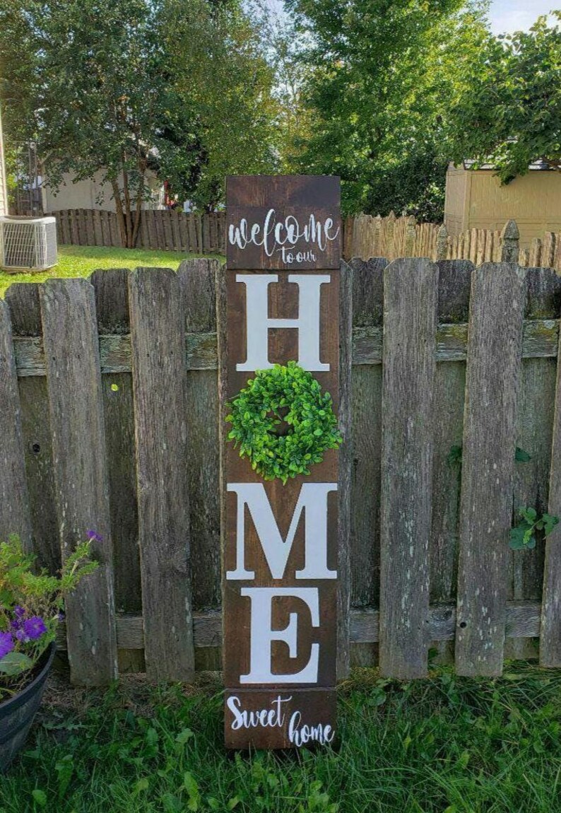May include: A wooden sign with the word "HOME" in large white letters. The letter "O" is replaced with a green wreath. The sign also includes the phrases "welcome to our" and "Sweet home" in cursive script. The sign is placed in a grassy yard.