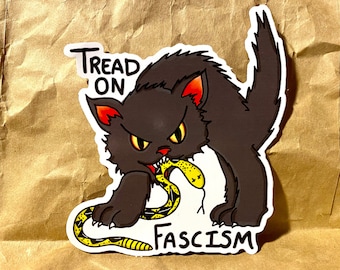 Puede incluir: Un gato negro con ojos rojos está mordiendo una serpiente amarilla con el texto "Tread On Fascism" debajo.