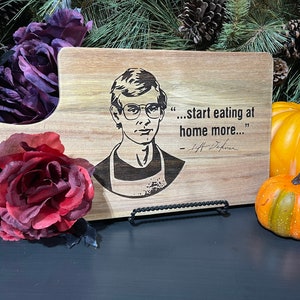 May include: A wooden cutting board with a black and white image of a man with glasses and the quote "...start eating at home more..." - Jeff Dahmer.