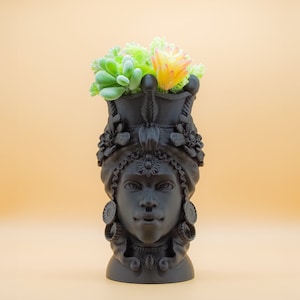 May include: A black decorative head-shaped vase with a detailed design, adorned with floral and ornamental elements. The vase holds a collection of artificial succulents in shades of green and yellow. The vase has a matte finish and is set against a light orange background.