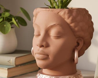 Peut inclure: Un pot de fleurs en forme de tête couleur terre cuite représentant un visage aux yeux fermés avec des cheveux texturés. Le pot repose sur une base en or rose métallique et porte des boucles d'oreilles créoles. Une plante succulente verte est plantée.