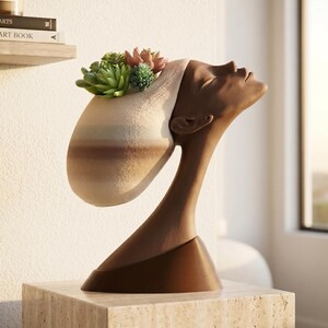 May include: A dark brown sculpture of a human head and neck in profile, tilted upward. The top of the head features a textured, light-colored planter section containing several green and pink succulent plants. The base is smooth and dark brown.