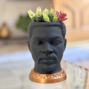 May include: A dark gray planter shaped like a man's head, with a bronze-colored base. The planter is filled with green and pink faux succulents. The head has detailed facial features and a beard.