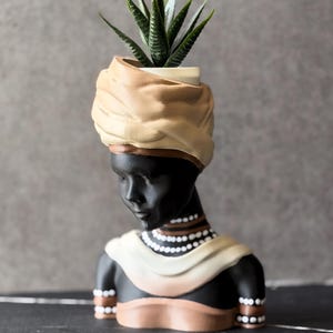 May include: A decorative planter shaped like a bust of a person with black skin, wearing a tan headwrap and a cream-colored shawl. The figure features white beaded necklaces and armbands, and holds a small green succulent plant in the top opening.