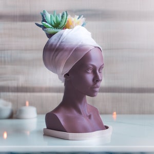 May include: A brown bust sculpture with a white turban and green and yellow succulents on top. The bust is on a white base and there are candles in the background.